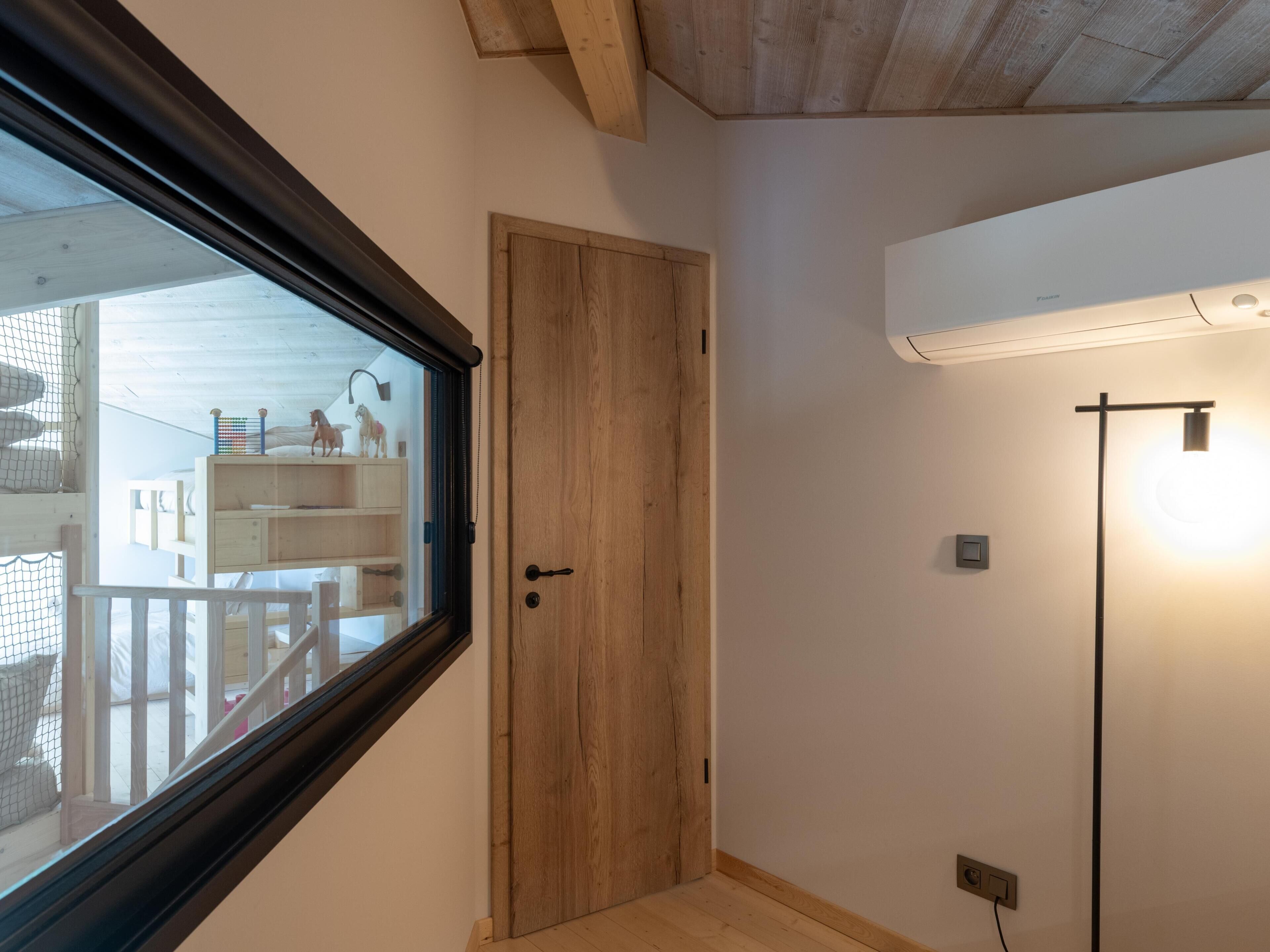 A room with a wooden door, a window with a view of a balcony, and an air conditioning unit on the wall.