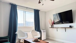 Ein modernes Wohnzimmer mit großem Fenster, einem Wand-Fernseher, einem Holztisch, zwei weißen Stühlen, einem grünen Sofa und einem Stehlüfter.