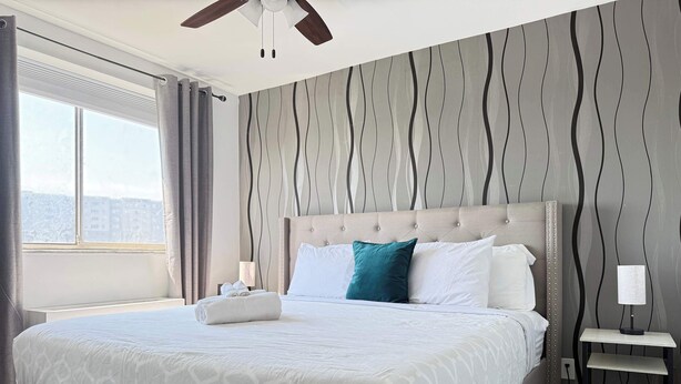 Ein Hotelzimmer mit einem großen Bett, einem Kopfteil, einem Nachttisch, einer Lampe und Blick auf die Stadt durch das Fenster.