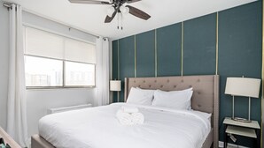 Una habitación de hotel con una cama grande, un ventilador de techo, una mesita de noche con lámpara y una ventana con persianas.