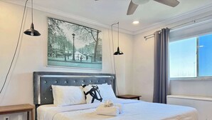 Une chambre d’hôtel moderne avec un grand lit, un ventilateur de plafond, un tableau représentant une rue de la ville et une fenêtre avec des rideaux.