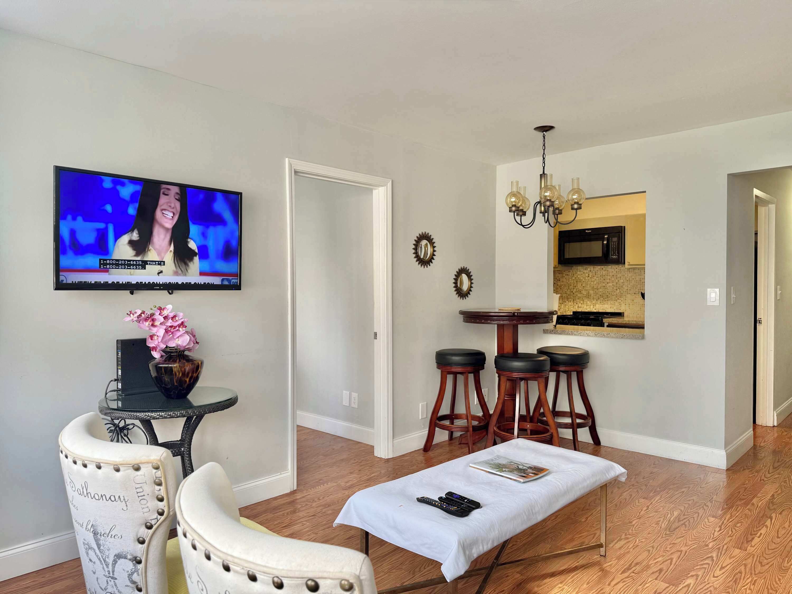 A living room with a television, a dining area with stools, a kitchen in the background, and a hallway leading to another room.