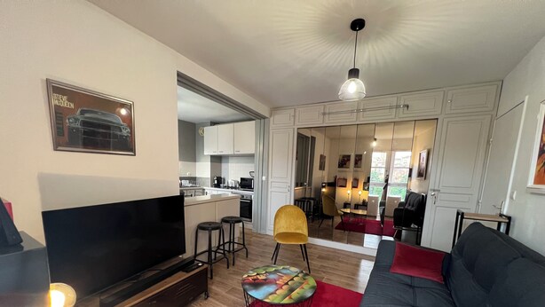 A modern living room with a flat-screen TV, a sofa, a red rug, and a dining area with a yellow chair. - Bright apartment overlooking the Tarn (Albi)