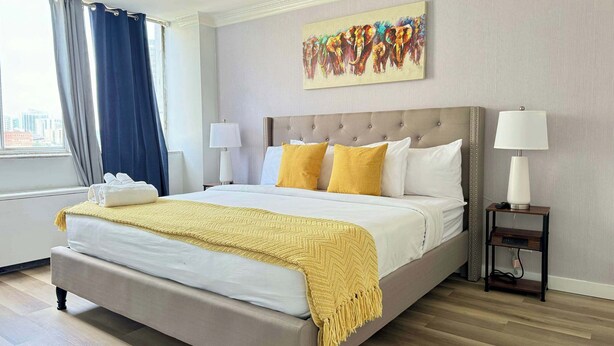 A hotel room with a large bed, two bedside lamps, a painting on the wall, and a view of the city through the window.