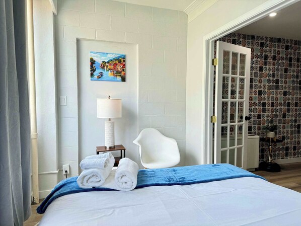Habitación de hotel con cama, una silla blanca, un cuadro de una escena costera, una lámpara y una puerta que da a un baño con azulejos decorativos.