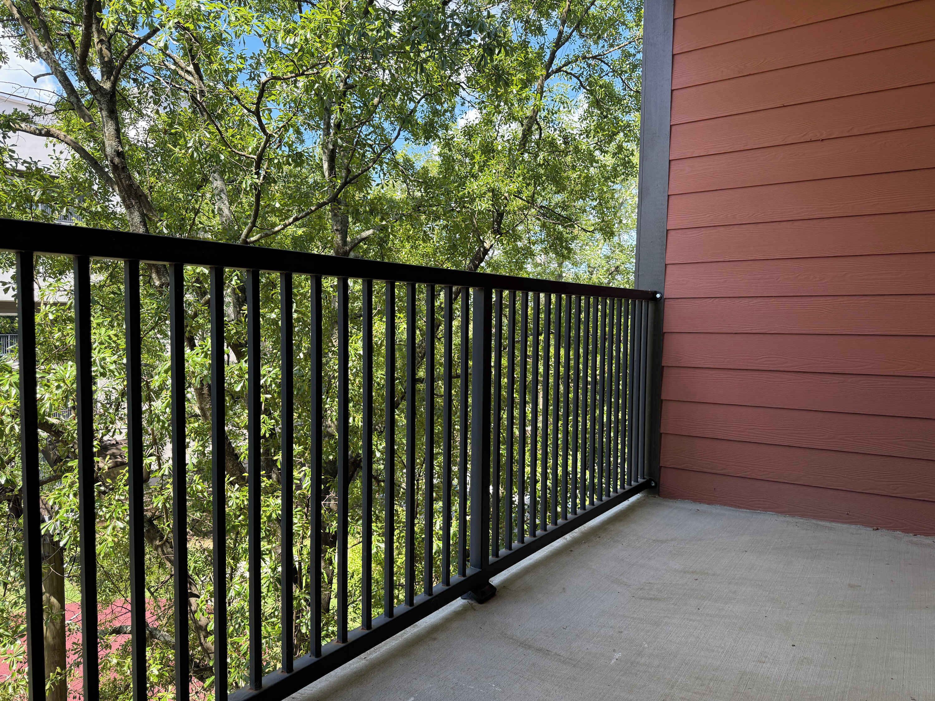 A balcony with a black metal railing, a concrete floor, and a view of green trees and a building in the background.