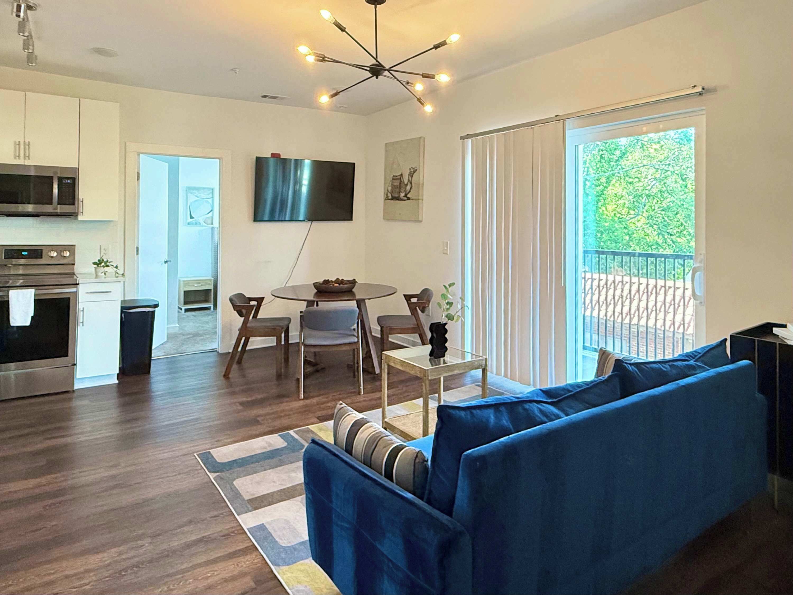 A modern living room with a blue sofa, a dining table with chairs, a flat-screen TV mounted on the wall, and a balcony with a view of greenery.