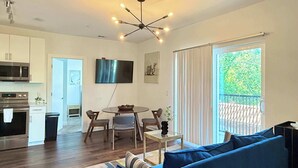 A modern living room with a blue sofa, a dining table with chairs, a flat-screen TV mounted on the wall, and a balcony with a view of greenery. - Upgraded Apartment in Old Fourth Ward w/ Patio (Atlanta)