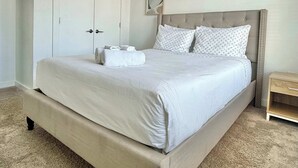 A neatly made bed with white linens and two rolled towels, a tufted headboard, a small bedside table, and a door with a handle. - Upgraded Apartment in Old Fourth Ward w/ Patio (Atlanta)