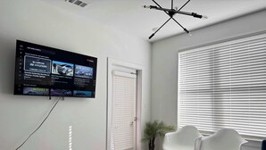 Een moderne woonkamer met een flatscreen-televisie aan de muur, waarop reclames voor auto's te zien zijn, een deur, een raam met zonwering, een kleine tafel met een afstandsbediening, en twee witte stoelen.