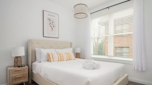 A neatly made bed with white bedding and a rolled towel, a framed picture on the wall, a bedside lamp, and a window with blinds.