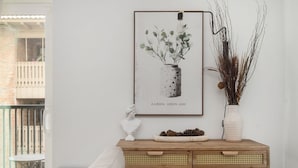 A room with a white wall, a framed artwork of a vase with green leaves, a white bust, and a wooden console table with a basket of pinecones.