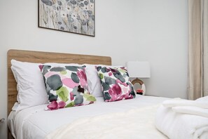 A bed with white bedding, two colorful pillows, a wooden headboard, a wall-mounted lamp, and a floral painting above the bed.