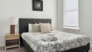 A bedroom with a bed, a nightstand, a lamp, a framed picture on the wall, and a window with blinds. - Downtown 1B1B Modern Apartment! Free Street Parking! (Atlanta)