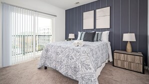 A bedroom with a large bed, two bedside tables, a window with blinds, and a view of buildings outside.