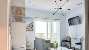 A modern living room with a grey sofa, a small round table with a vase, a TV mounted on the wall, and a window with blinds.