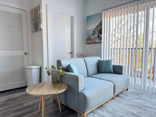 Un salon moderne avec un canapé gris, une table ronde en bois et une fenêtre équipée de stores verticaux.