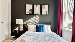 A neatly made bed with a patterned bedspread, a bedside lamp, and a nightstand. There are three framed artworks on the wall above the bed.