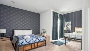 Habitación de hotel moderna con una cama grande, un escritorio y una silla. Hay una mesita con una lámpara y un marco de fotos. La habitación tiene un papel pintado y una ventana con cortinas.
