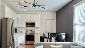 Eine moderne Küche mit Edelstahlgeräten, ein Essbereich mit einem schwarzen Tisch und Stühlen sowie ein graues Sofa mit Kissen.