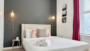 A neatly made bed with white linens and rolled towels, a small bedside table with a lamp, and a window with red curtains. - Modern Multi-level 2B2B Apartment in Old Fourth Ward (Atlanta)