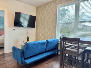 Un salon avec un canapé bleu, un téléviseur fixé au mur, une table à manger avec des chaises et une fenêtre avec des stores.
