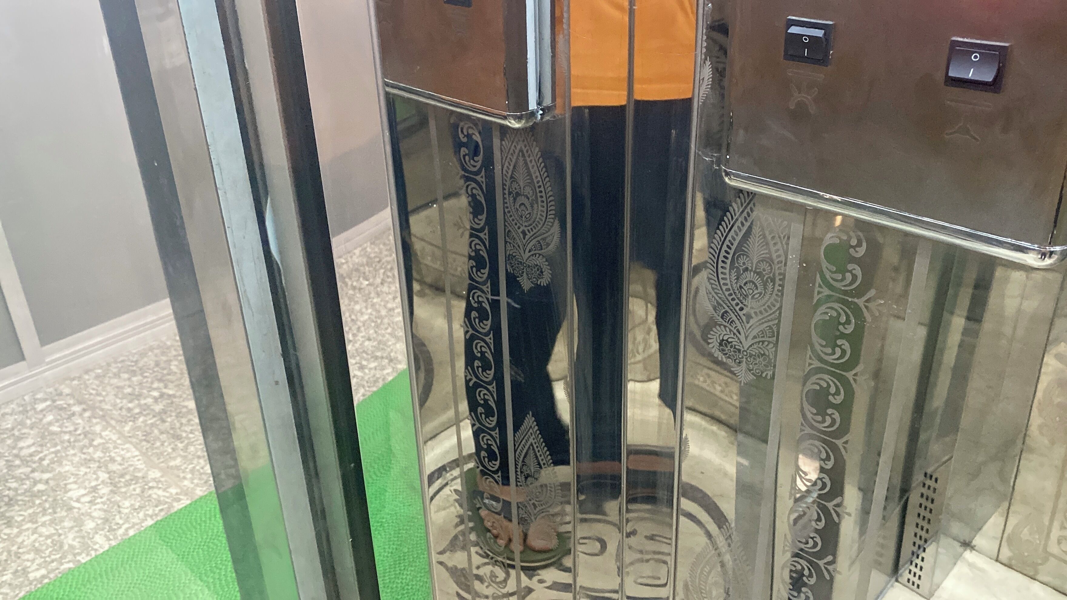 Un ascensor con paredes metálicas reflectantes y piso cubierto de moqueta verde.