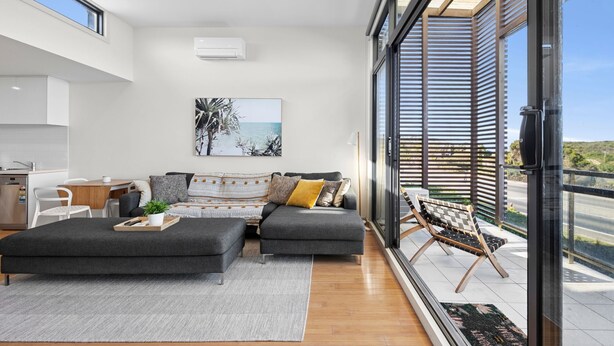 A modern living room with a grey sofa, a black ottoman, and a wooden coffee table. There is a large window with a view of the sea and a balcony with a chair. - Inlet Apartment I Walk to beach, cafes and more (Aireys Inlet)