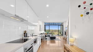 A modern kitchen with white cabinets, a large window, and a dining area with a wooden table and chairs. - Inlet Apartment I Walk to beach, cafes and more (Aireys Inlet)