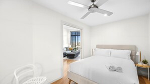 A modern bedroom with a bed, a ceiling fan, a nightstand, and a view of a living area through an open door. - Inlet Apartment I Walk to beach, cafes and more (Aireys Inlet)