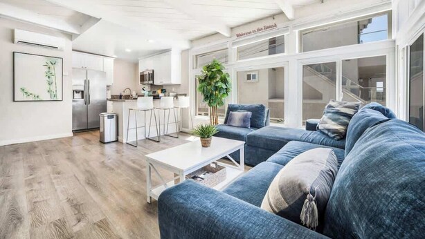 A modern living room with a blue sofa, a white coffee table, and a kitchen area in the background. - Boho Bliss | Mission Beach Retreat w/ Surfboards (San Diego)