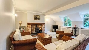A spacious living room with a fireplace, comfortable seating, a television, and a view of greenery through large windows.