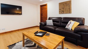 A living room with a wooden coffee table, a leather sofa, a flat-screen TV, and a wooden door. - 3 bedroom accommodation in near Gatehouse of Fleet (near Gatehouse of Fleet)