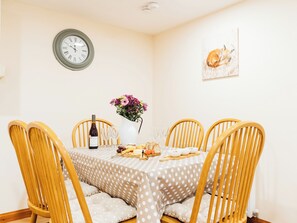 A dining area with a round table, four wooden chairs, a vase of flowers, a wine bottle, and a clock on the wall.