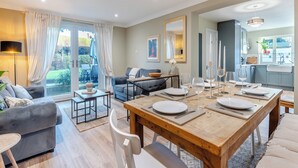 Una sala de estar moderna con un sofá gris, una mesa de comedor de madera lista para cuatro comensales y una cocina al fondo.