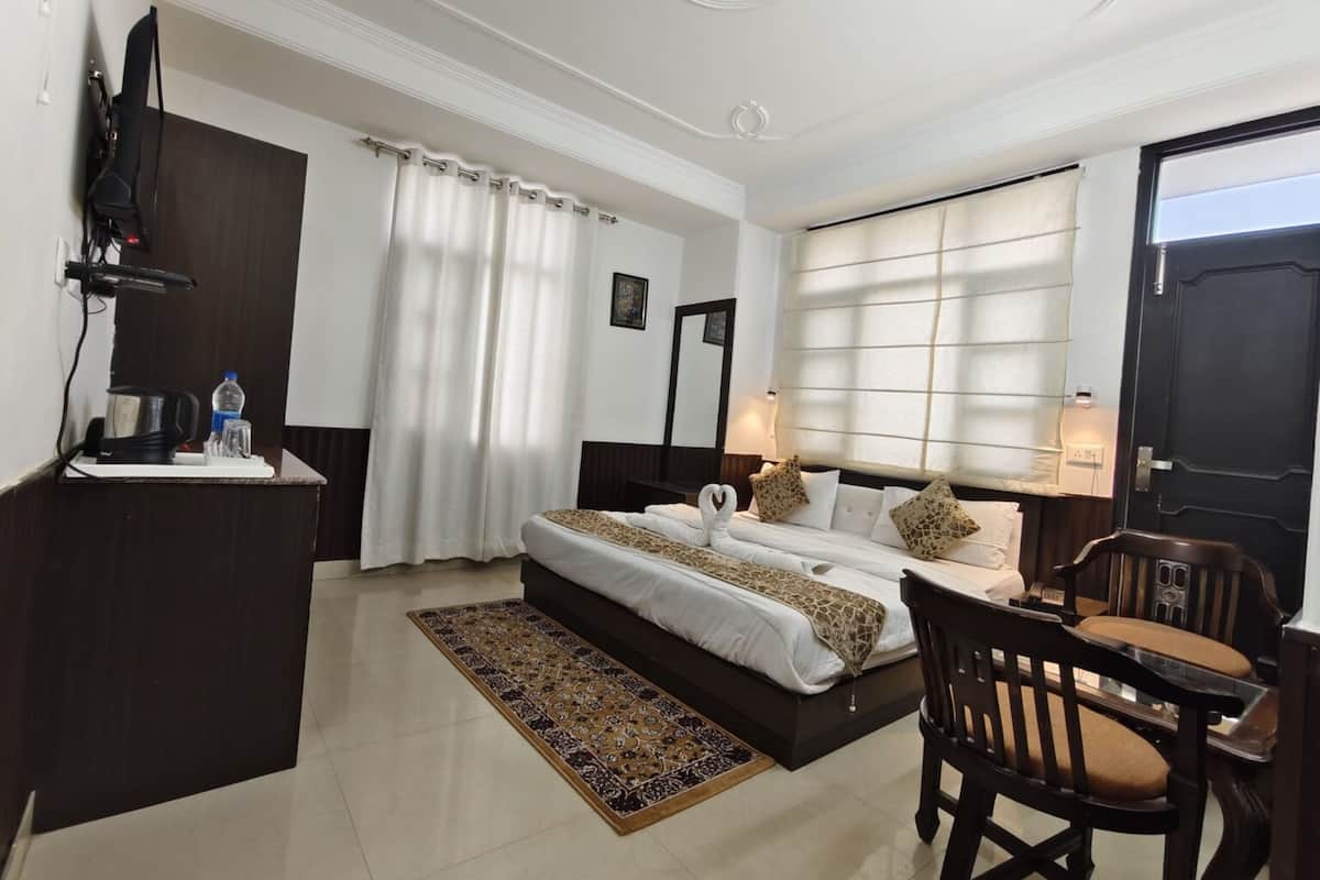 A hotel room with a bed, a desk with a kettle, a chair, and a window with curtains.