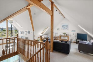 A spacious loft with a high ceiling, wooden beams, a staircase, a living area with a sofa, a TV, and a view of the outside.