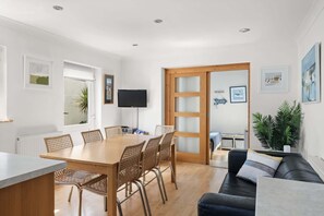 A modern living room with a wooden dining table, wicker chairs, a black leather sofa, and a TV mounted on the wall.