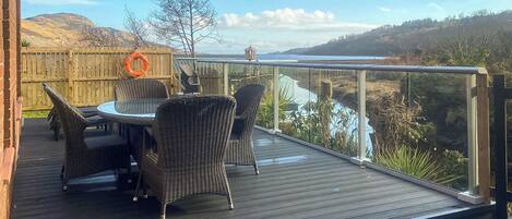 A deck with wicker furniture and a glass railing overlooking a river.