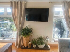 Sala de estar con un televisor de pantalla plana montado en la pared, una planta grande en maceta, un pequeño farol y vista a un balcón con barandilla.