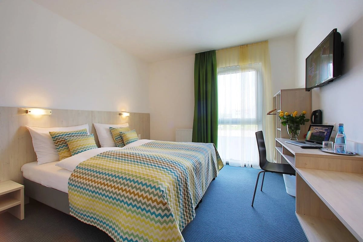 Une chambre d’hôtel avec un lit, un bureau, une chaise, une télévision et une fenêtre avec des rideaux.