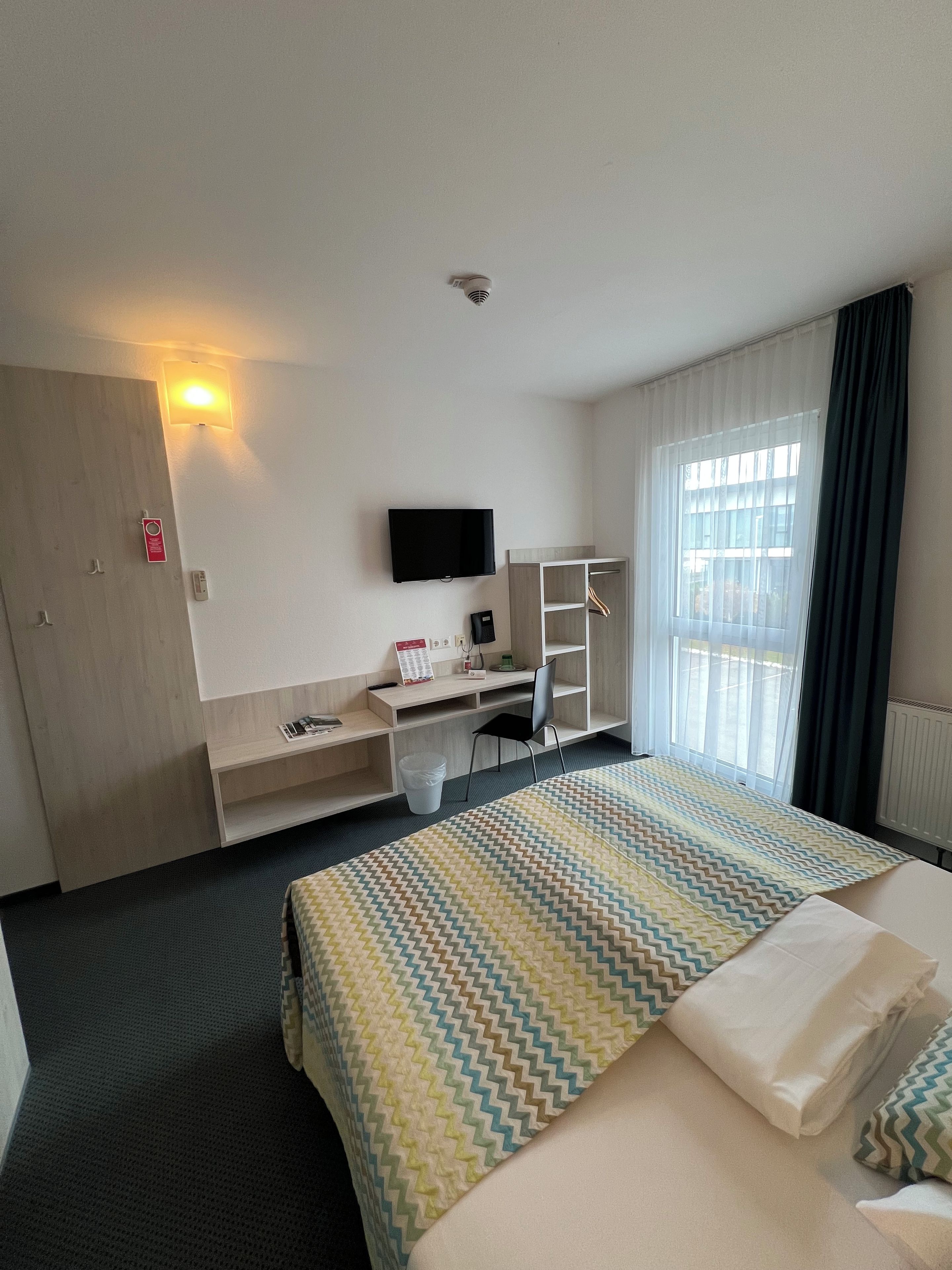Une chambre d’hôtel avec un lit, un bureau avec un ordinateur, une télévision et une fenêtre avec des rideaux.