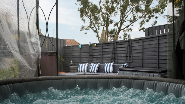 A hot tub with a view of a patio area, including a sofa with striped cushions and a wooden fence.