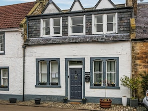 Une maison traditionnelle à deux étages, avec une façade blanche, un toit foncé et une porte bleue. Des jardinières sont posées sur les rebords des fenêtres, et un pot de fleurs se trouve dans le jardin.
