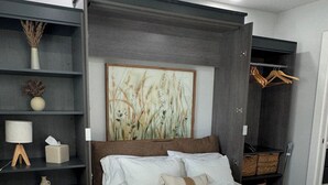 A modern bedroom with a large bed, a dark headboard, a wall-mounted shelf with decorative items, and a painting above the bed. - Charming 1-bedroom Guesthouse in Central Austin (Austin)