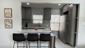 A modern kitchen with a central island, three black bar-style chairs, stainless steel appliances, and a window with blinds. - Charming 1-bedroom Guesthouse in Central Austin (Austin)