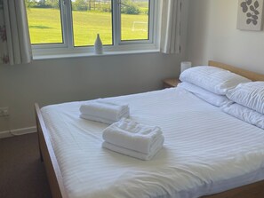 Een netjes opgemaakt bed met witte lakens, een raam met uitzicht op een groen veld en een nachtkastje met een lamp.