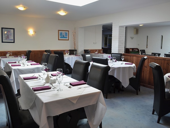 Un restaurant doté de nombreuses tables, chacune recouverte d’une nappe blanche, agrémentée de chaises noires et de couverts en argent.