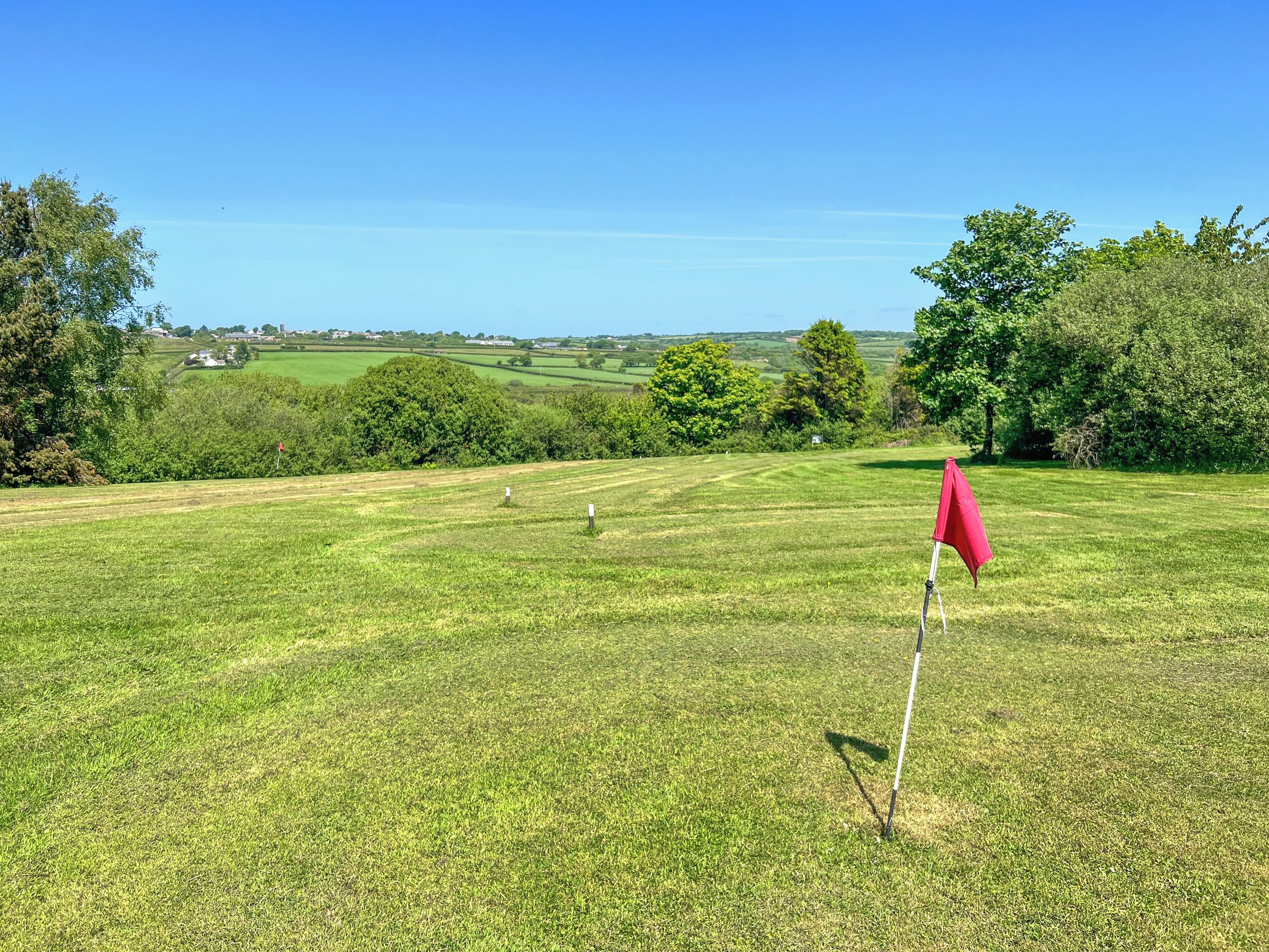 Een golfbaan met een rode vlag, groen gras, bomen en gebouwen in de verte.