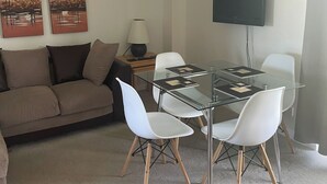 Un salon moderne avec une table à manger à plateau de verre, des chaises blanches, un canapé marron, une lampe et un téléviseur à écran plat fixé au mur.